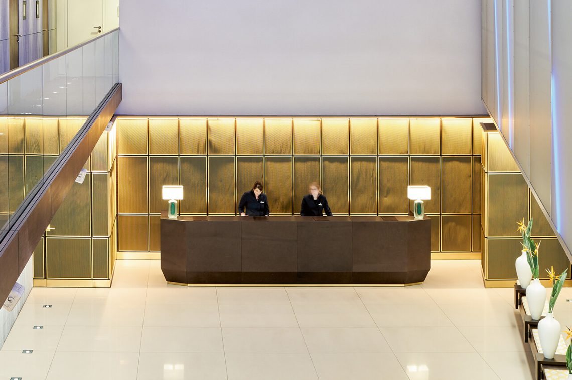 The SIDE Hotel reception desk in front of the backlit brass wall, with two cylindrical table lamps