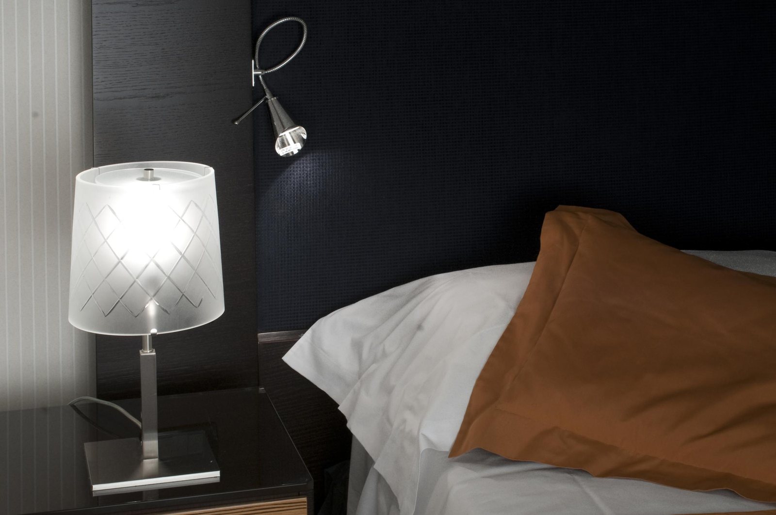 The bedside table with the etched glass lamp and the flexible arm aimed at the pillow