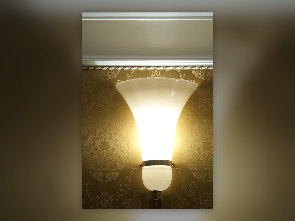 The trumpet wall light in opal glass, lit against the gold damask wall