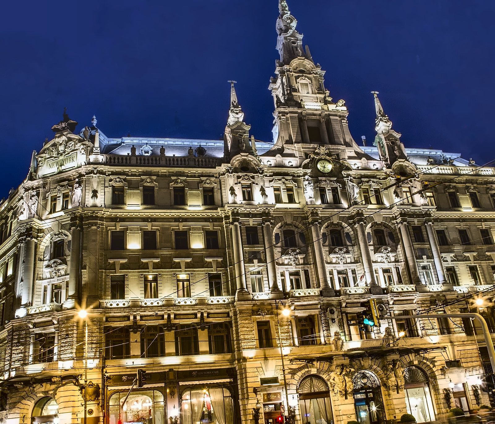 Die Fassade des New York Palace bei Nacht, von unten gesehen: Säulen, Gesimse und Statuen beleuchtet bis hinauf zum Turm, und Lichtspuren auf der Straße