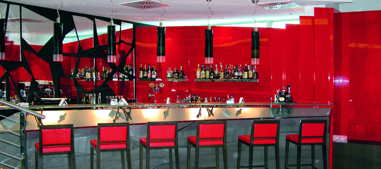 Die Bar des UNA Hotel Malpensa: eine Reihe schwarzer zylindrischer Pendelleuchten aus Muranoglas über der Metalltheke, zwischen roter Wand und roten Barhockern
