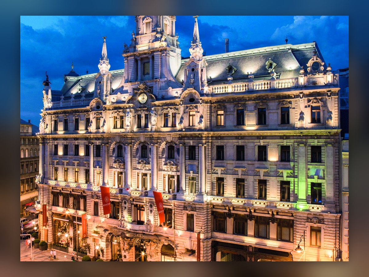 Die Fassade des New York Palace in Budapest in der Dämmerung, von oben gesehen: beleuchtete Gesimse, Balkone und Statuen unter blauem Himmel