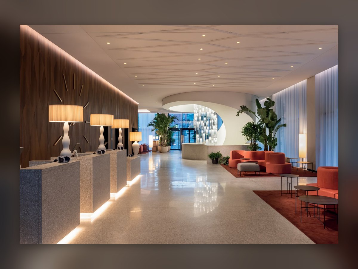 The lobby of the Mövenpick Hotel Basel: table lamps with a sinuous white base and a lit pleated shade on the reception desks, coral sofas on the right