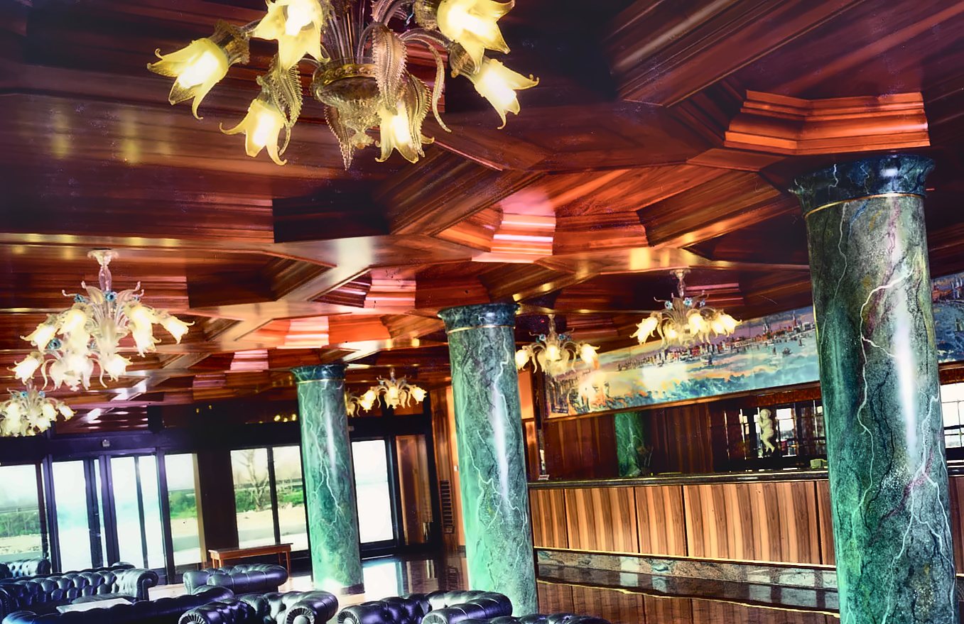Die Lobby des Russott Hotel in Mestre: leuchtende strohgelbe venezianische Kronleuchter mit Blättern und Blüten unter der hölzernen Kassettendecke, grün geäderte Säulen und schwarze Ledersessel