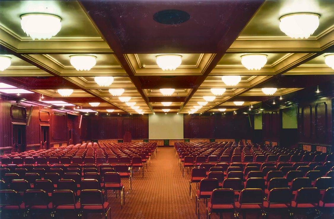 Der Kongresssaal des Russott Hotel in Mestre: runde Deckenleuchten aus Muranoglas leuchten in den hellen Deckenkassetten, Reihen roter Stühle und holzvertäfelte Wände