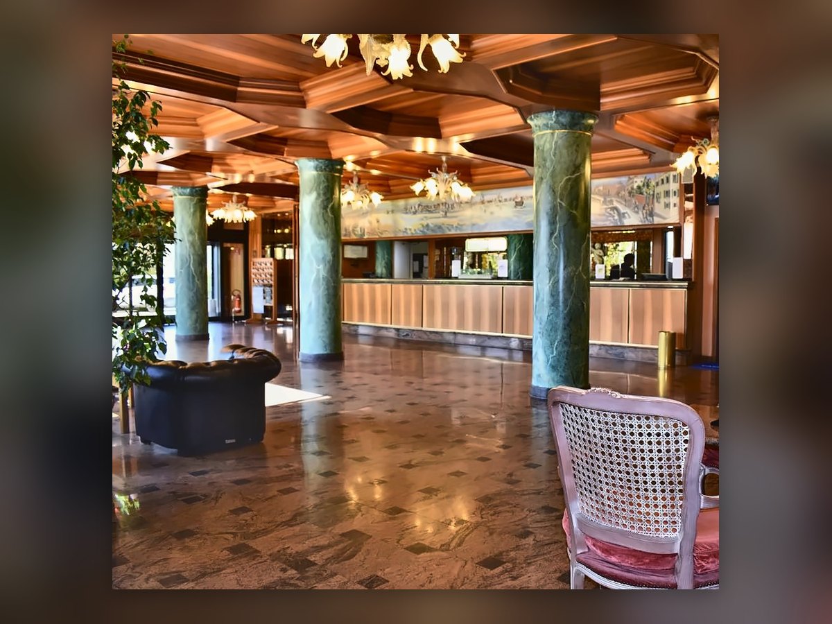 Die Lobby des Russott Hotel in Mestre mit leuchtenden venezianischen Kronleuchtern über rosa Stühlen mit Wiener Geflecht und den grünen Säulen, im Hintergrund die Rezeption