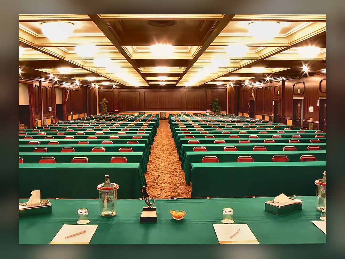 Der Kongresssaal des Russott Hotel, bestuhlt für eine Tagung: runde Deckenleuchten leuchten in den Kassetten, Reihen grün bespannter Tische und rote Stühle