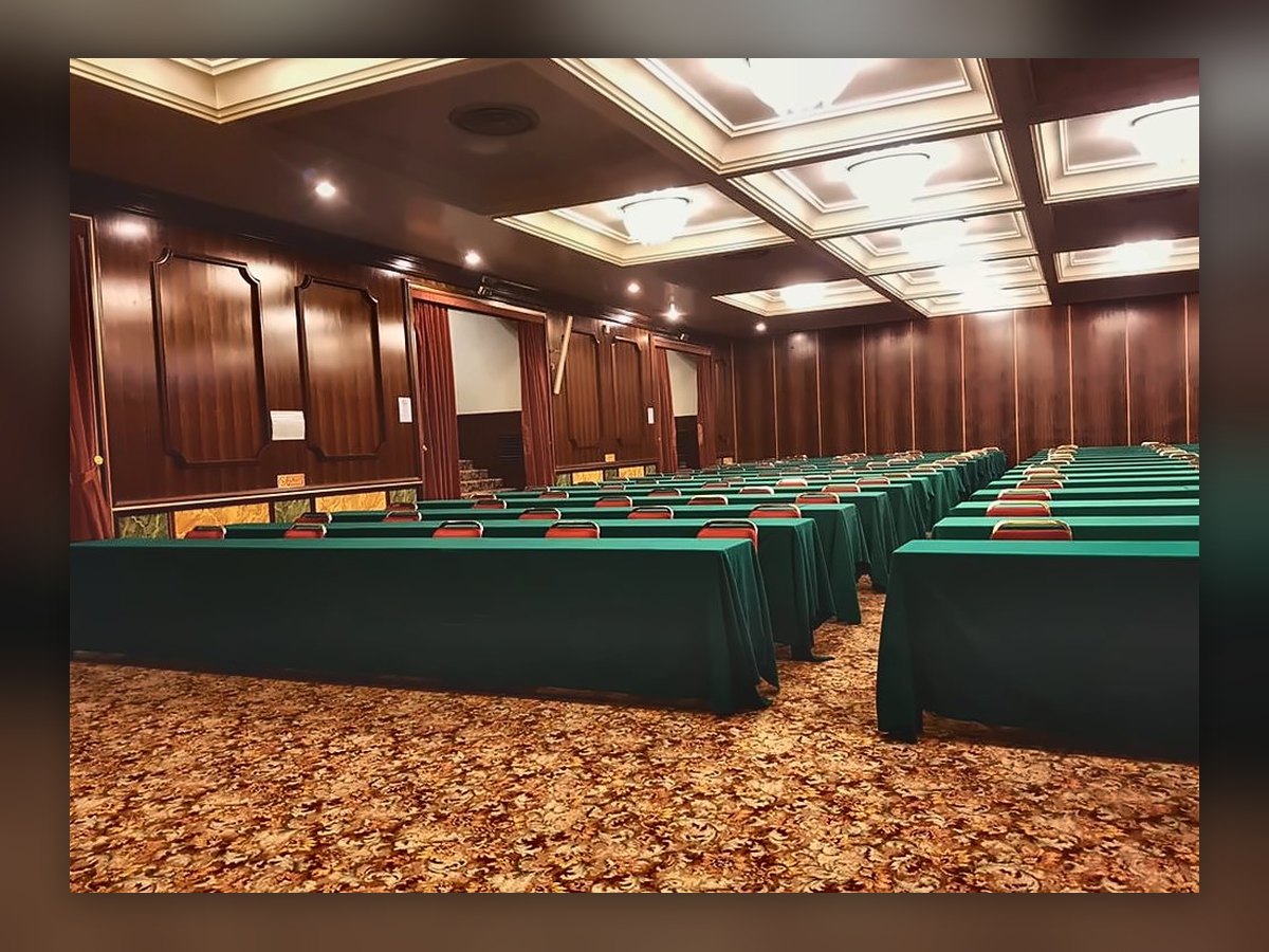 Ein Saal des Kongresszentrums im Russott Hotel von der Seite: runde Deckenleuchten in den Deckenkassetten, grüne Tische in Reihen und Holzwände mit roten Vorhängen
