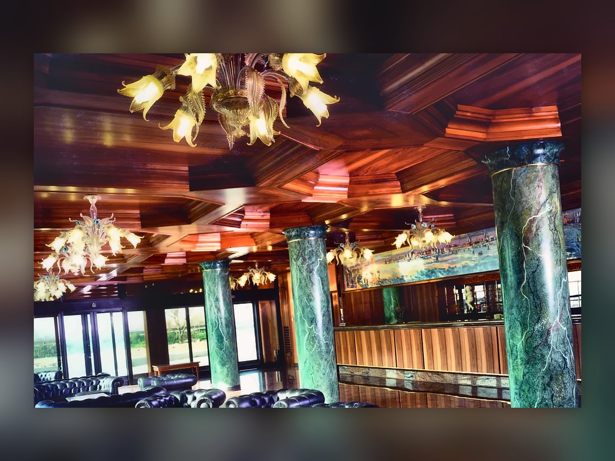 The lobby of the Russott Hotel in Mestre: lit straw-coloured Venetian chandeliers with leaves and flowers under the coffered wooden ceiling, veined green columns and black leather armchairs